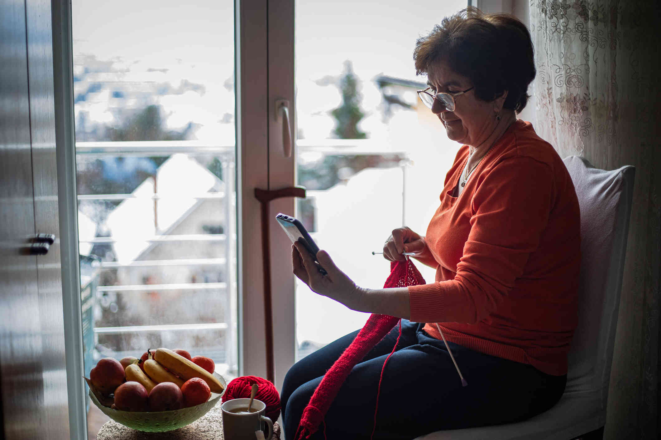 A middle aged woman in a red shirt sits in her home knitting while looking at the phone in her hand.